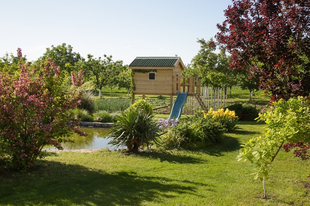 Domek ogrodowy dla dzieci postawiony na słupach, z zamontowaną zjeżdżalnią. 